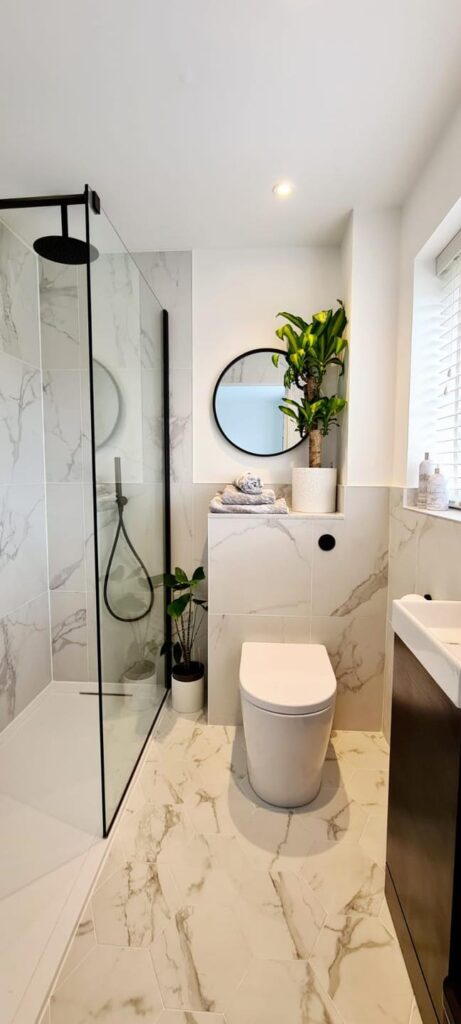 Modern bathroom interior with white tiling, shower, toilet, and sink, featuring marble-effect floor and walls, and green plants.