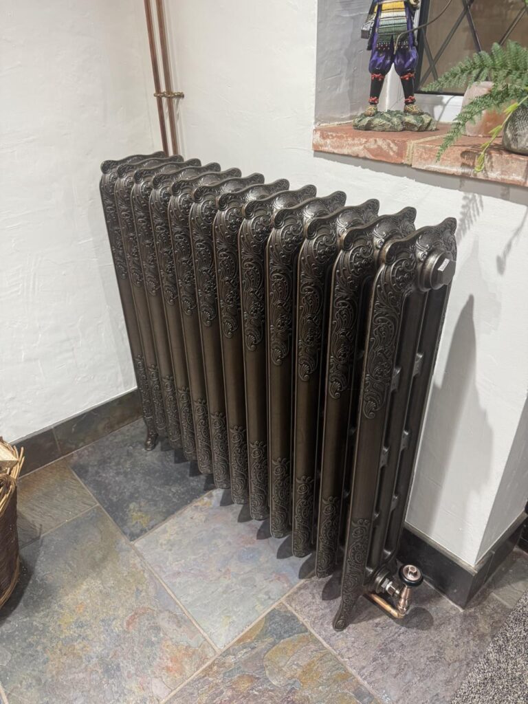 A dark bronze radiator with ornate details stands against a white wall in a room with grey tiling and copper plumbing.