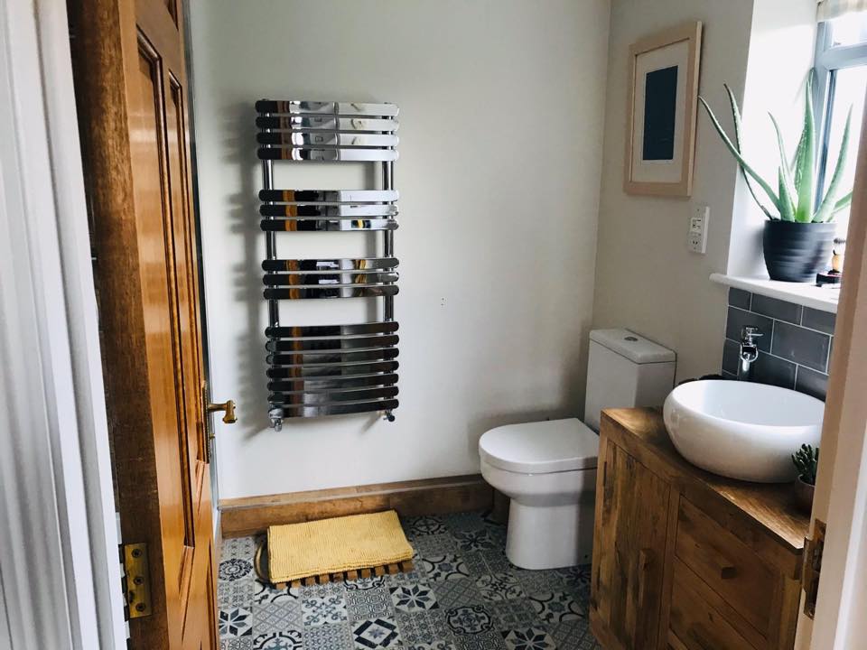 A bathroom interior with white walls, wooden accents, and patterned tiling on the floor, featuring a toilet, sink, and heated towel rail near plumbing fixtures.