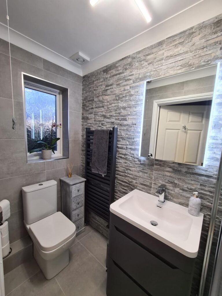 Modern bathroom interior with grey tiling, toilet, sink, and shower area, featuring white and dark grey fixtures, creating a clean and minimalist atmosphere.