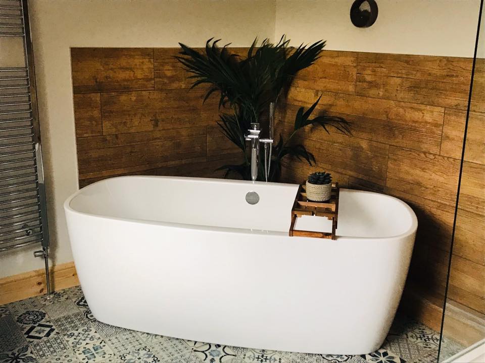 A freestanding white bathtub with silver plumbing, wooden accents, and decorative tiling in a bathroom setting with a plant and shower